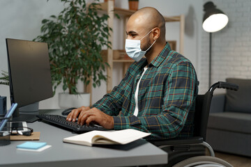Young disabled man in wheelchair sitting at his working table wearing face mask