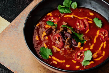 spicy tomato soup with beans, chili pepper, cheese and parsley in a black plate on a copper background