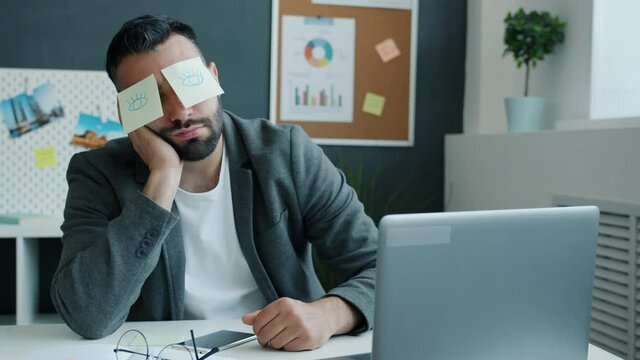 Tired businessman napping in office sitting at desk having sticky notes with open eyes pictures covering face while guy relaxing during work break.