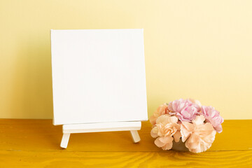 Blank white canvas with carnation flowers on wooden table. yellow background