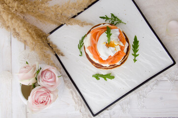 Pancakes with salmon, poached egg and arugula on white plate on a light background. Healthy breakfast. 