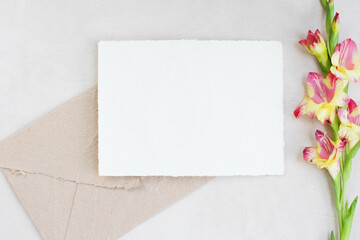 Photostock composition. Feminine desktop mockup scene with leaves, Amaryllis flower, silk ribbon, brown envelope, blank greeting card on white textured concrete background. Flat lay, top view