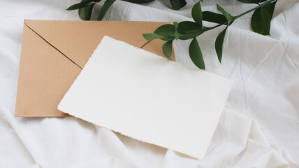 Photostock wedding styled composition. Feminine envelope mockup scene with ruscus leaves, silk ribbon, blank greeting card, on creme textured fabric background. Flat lay, top view.