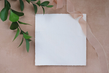 Summer wedding stationery mock-up scene. Blank card, invitation in sunlight. Ruscus leaves, greeting card and silk ribbon. Concrete brown texture background. Flat lay, top view.
