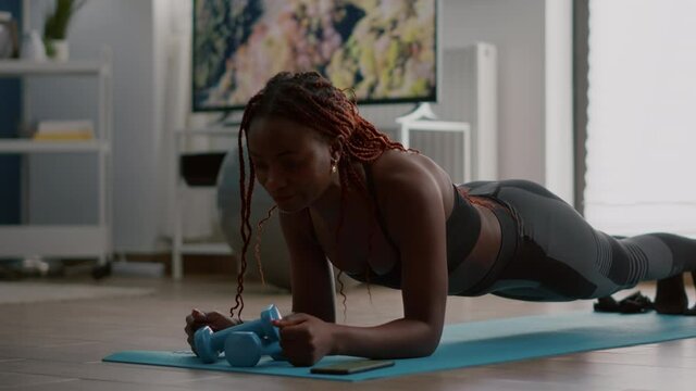 Athlete african in sportwear doing fitness exercices putting cronometer on phone sitting on yoga map in living room. Flexible woman stretching muscle body enjoying healthy lifestyle