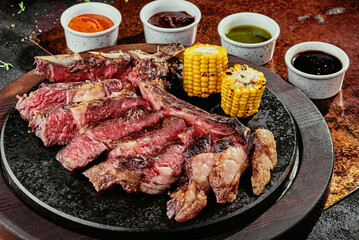 T-Bone Steak serving with grilled vegetables and various sauces on a black round plate on a background of greens, spices and vegetables, on a copper sheet.