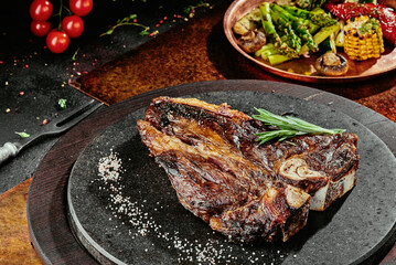 T-Bone steak serving with grilled vegetables on a black round plate on a background of greens,...