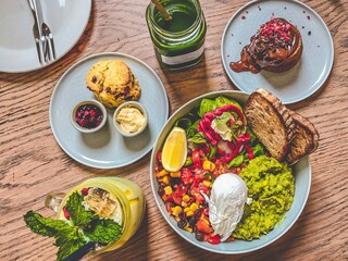 a healthy bowl of mixed vegetable salad and fresh vegetable juice and smoothie on a wooden table accompanied by pastries, surrounded by clean plates and cutlery. Food photography.
