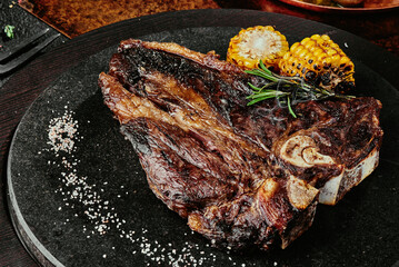 T-Bone steak serving with grilled vegetables on a black round plate on a background of greens,...