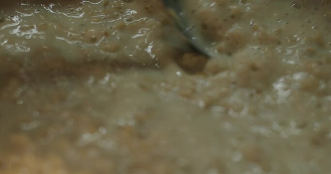 The cook is stirring the boiling oatmeal milk porridge with a tablespoon