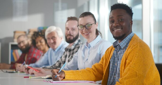 Multiethnic adult people sitting in row at desk and smiling at camera in classroom
