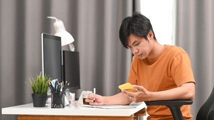 Young man freelancer using smart phone and making note on notebook.