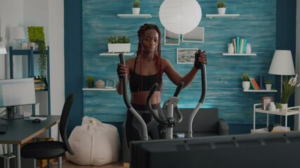 Black woman athletic training on elliptical bike practicing cardio sport watching aerobic video on tv during bodyweight morning routine in living room. Trainer enjoying healthy lifestyle