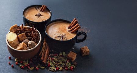 Masala tea traditional hot Indian sweet milk spiced drink in black cups. Spices cinnamon sticks, cardamom pods, star anise and coriander seeds in brown ceramic cup on a black slate stone plate