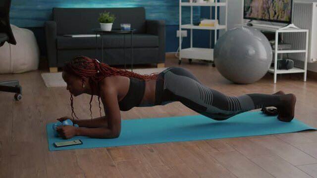 Flexible slim black woman stretching body muscles putting cronometer on phone practicing sport on yoga map during fitness workout in living room. Athetic adult enjoying healthy lifestyle