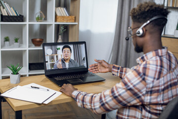 Back view of afro american man in headset sitting at home and having video conversation on laptop with asian coworker. Concept of pandemic time and distance cooperation.