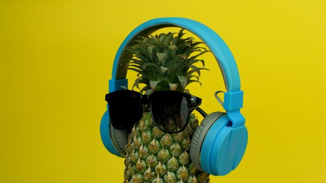 Whole unpeeled pineapple with sunglasses and headphone listening to music. Closeup shot of ripe juicy pineapple / Ananas with stalk rotating slowly against a yellow background - summer fruit concept