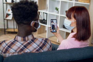 Multiracial couple in face mask sitting on couch and having video conversation with muslim friends. Diverse friends using modern gadgets for distance communication during pandemic.