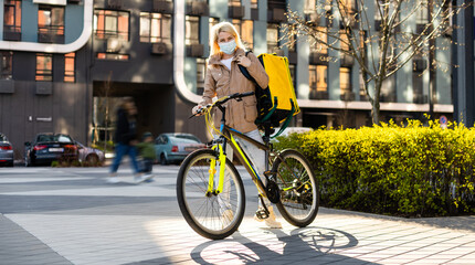 Fototapeta premium Portrait of beautiful woman delivery worker in medical mask for riding bike standing at street. Pretty female courier with backpack. Food delivery concept