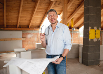 male architect with black glasses and blue shirt examines building plan on construction site in...