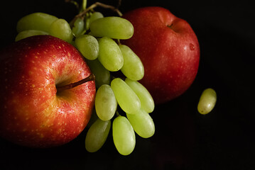 Two ripe fresh red apples with water drops on glossy peel and bunch of green grapes on black background