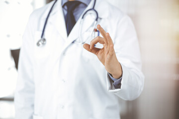 Unknown male doctor standing straight with OK sign in sunny clinic, closeup. Perfect medical service in hospital. Medicine and healthcare concept