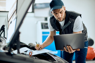 Auto repairman using laptop and writing data while running car diagnostic in a workshop.