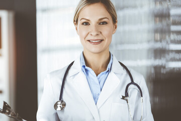 Friendly female doctor standing and holding clipboard in clinic . Portrait of cheerful smiling physician. Perfect medical service in hospital. Medicine and healthcare concept