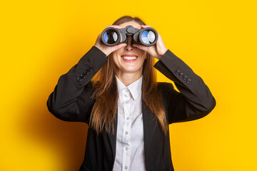 smiling young business woman looking through binoculars on yellow background - obrazy, fototapety, plakaty