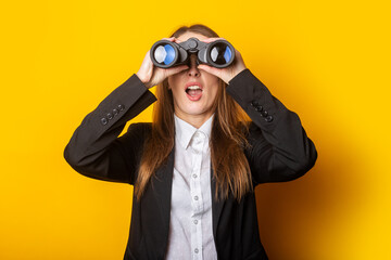 surprised young business woman looking through binoculars on yellow background - obrazy, fototapety, plakaty