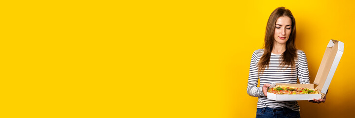 young woman looks thoughtfully at the pizza in the package on a yellow background. Banner