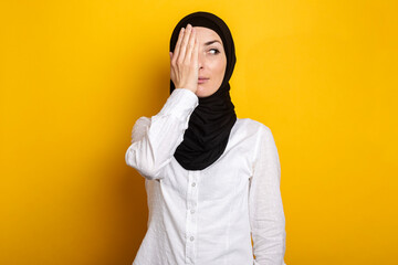 Young Muslim woman in hijab covering one eye with her hand smiles and looks to the side on a yellow background. Banner