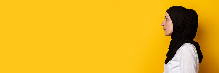 Young Muslim woman in hijab looks to the side on a yellow background. Banner. Profile shot, side view