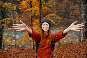 happy woman with a backpack travels in the park in autumn and gesticulate with the hands of the river in the background