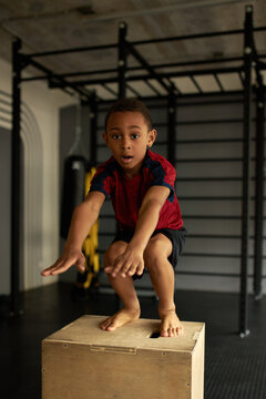 Plyometric training. Vertical image of active little dark skinned boy standing barefooted on plyo box doing jumps or sit ups to increase muscle strength. Athletic black child exercising in gym