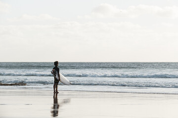 Surfer standing at the beach shore at sunset watching the waves