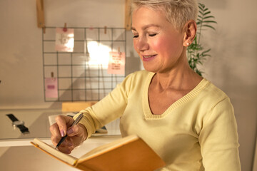 Successful happy mature woman posing in casual interior holding notebook and pen smiling feeling satisfied while crossing out accomplished goals, making to do list. Daily planning concept