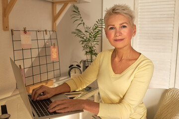 Indoor image of elegant short haired middle aged female teacher sitting at table in front of open laptop working remotely, having online distant class. Technology, job and occupation concept