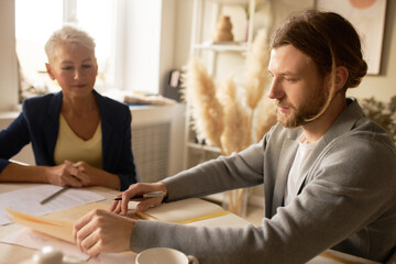 Portrait of attractive mature female teacher having individual class with her concentrated young bearded male student checking homework, preparing for exam. People, job and occupation concept