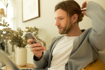 Focused fashionable young red haired guy with beard reading text message on generic mobile phone, having thoughtful pensive look while working on laptop. Technology, gadgets and connection