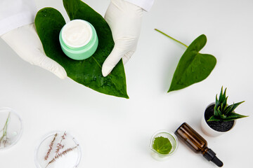 gloved hands of female lab technician hold Luxury skincare moisturizing cream in green glass jar on large tropical plant leaf. surrounded by symbols of natural cosmetics on backdrop of cosmetology lab