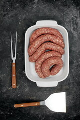 Sausages For Frying or BBQ Grilling In White Ceramic Dish And Grill Tool On Shabby Black Background, Top View. Raw Lamb Or Beef Sausages In Natural Casing In White Tray, Overhead View.