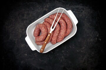 Sausages For Frying or BBQ Grilling In White Ceramic Dish And Grill Tool On Shabby Black Background, Top View. Raw Lamb Or Beef Sausages In Natural Casing In White Tray, Overhead View.