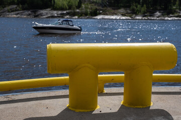 Closeup of a metallic bollard on the port with a yacht sailing across the horizon in the background.