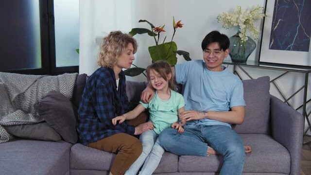 Multiracial family portrait on couch. Dad and daughter with felt-tip pictures on hands. Young asian father and caucasian mother with kid having fun in living room. Slow motion 4k