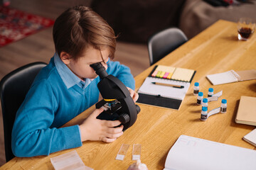 Distance online education, internet learning. First grade boy studying at home using microscope, making notes, biology online lesson