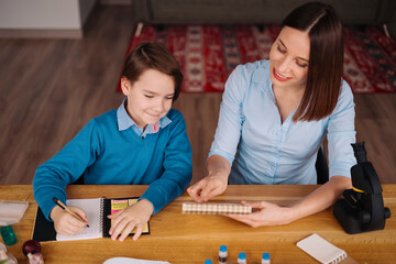 Fototapeta premium Aunt and nephew do homework together. Women explain kid writes in notebook. Schoolboy studying at home. Family relationships, primary education, childhood
