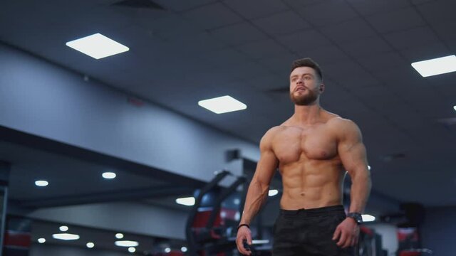 Sportsman with perfect muscular body in the gym. Young bodybuilder with naked torso stands in the gym and bothering his muscles. Sportive lifestyle.