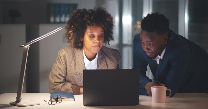 Young afro colleagues sitting together at desk working late at night in dark office on computer