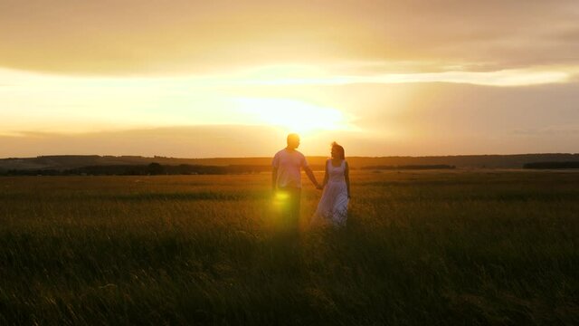 Silhouette of man and a woman at sunset. Romantic date and love in nature. A beautiful girl and a guy are walking hand in hand in field, in sun on the green grass. A young couple in love is traveling.